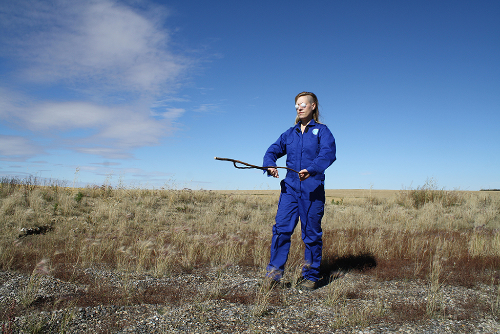 a person in blue coveralls with a divining rod standing in a field with a blue sky