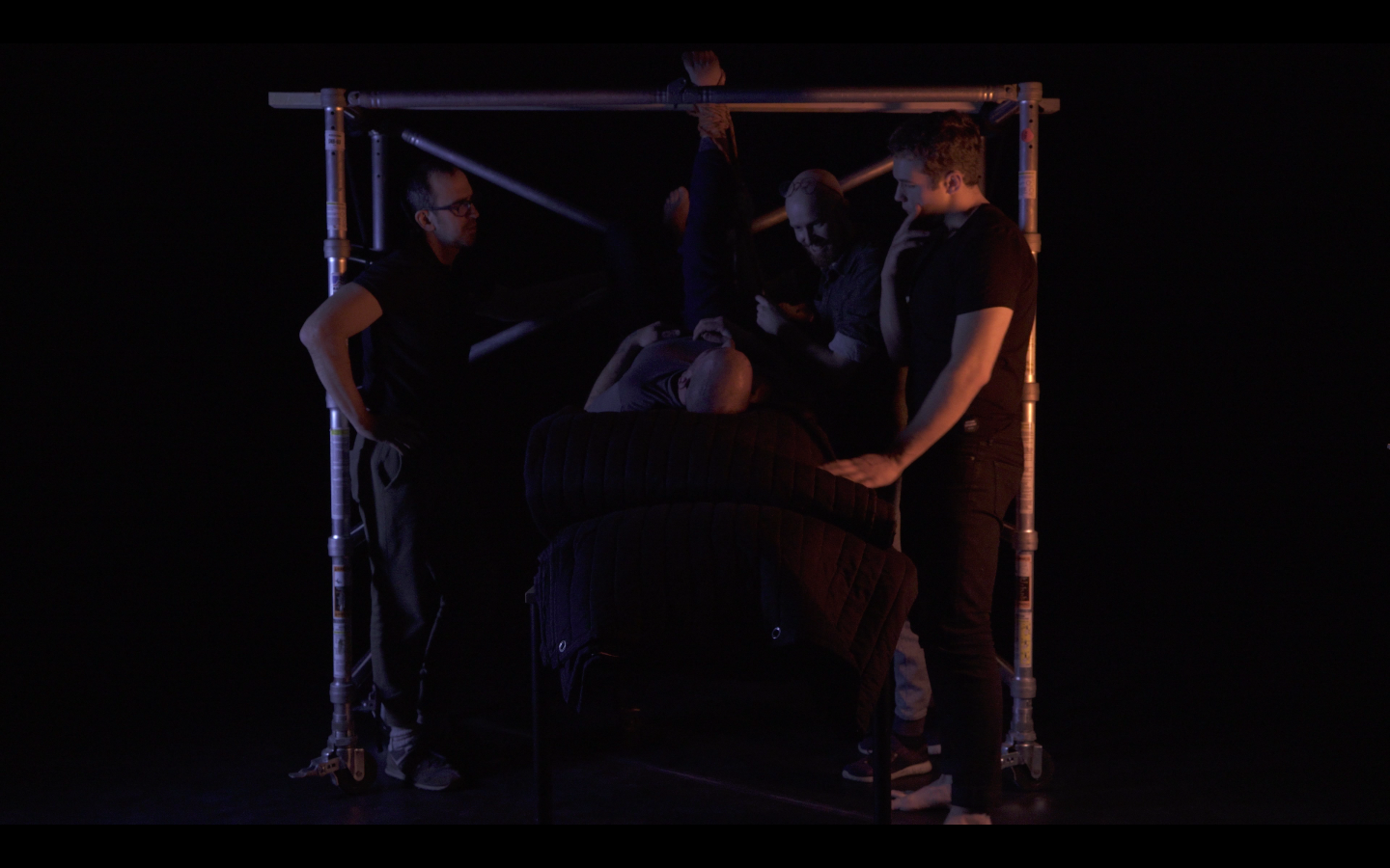 Dark background with several figures standing close together inside a scaffolding like structure, one figure appears to be preparing to hang upside down