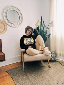 Cassandra lounging on a chair in a neutrally decorated room with a plant, soft light is coming in the window.